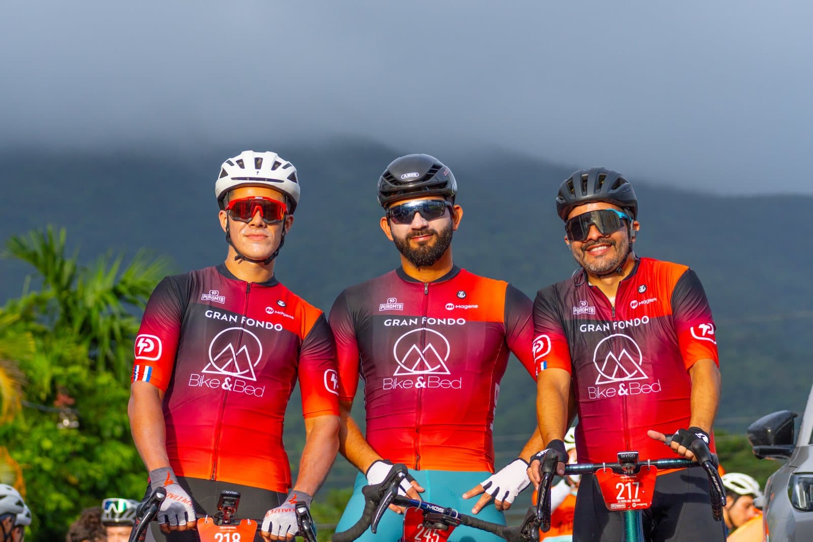 Tres ciclistas con el maillot de Bike & Bed sonriendo, con la montaña detrás